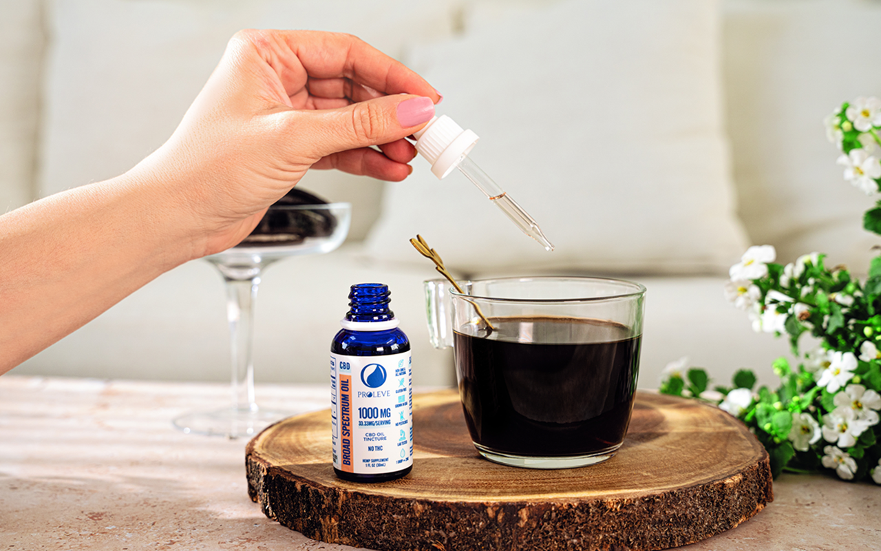 CBD tincture bottle open with hand holding tincture dropping cbd into a cup of coffee